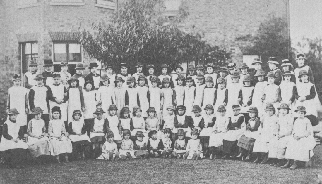 30-40 young girls of different ages are wearing white aprons and hats seated for a group photo outdoors.