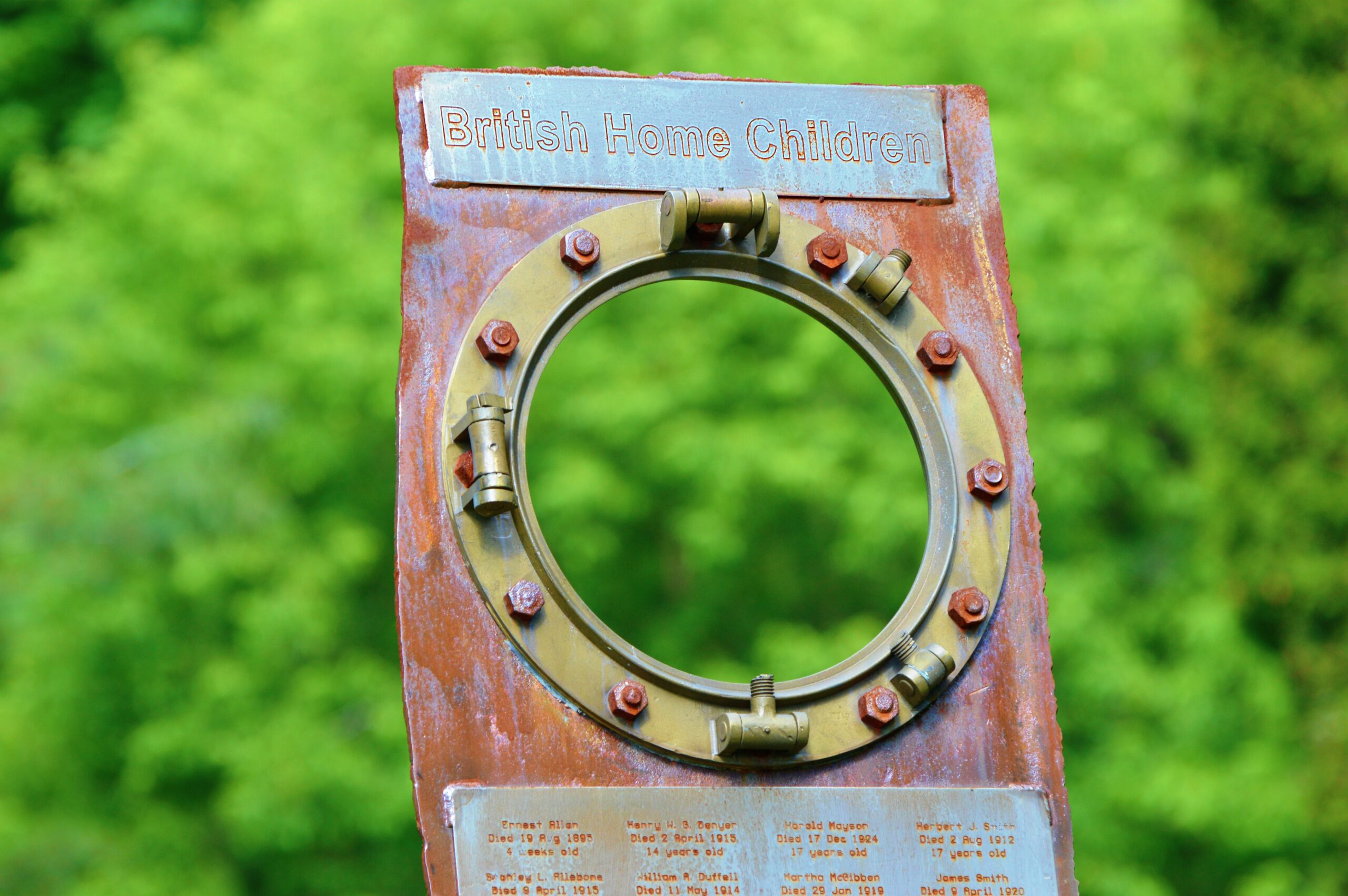 A small wooden and metal monument that reads, “British Home Children.” The monument is built from a rusted port hole from an old ship.