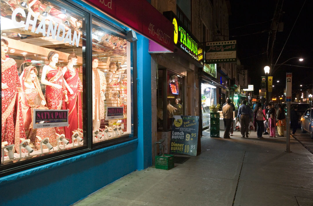 Photo faces down the sidewalk of Gerrard St E. showing the shop fronts. There is a shop with traditional South-Asian clothing in the display window with the name "Chandan Fashion". In the background a group of people crowd around the entrance to a restaurant.