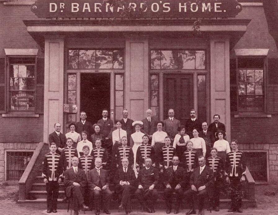 A group of staff and a visiting musical group stand outside a building. A sign on the building reads, “Dr. Barnardo’s Home.”