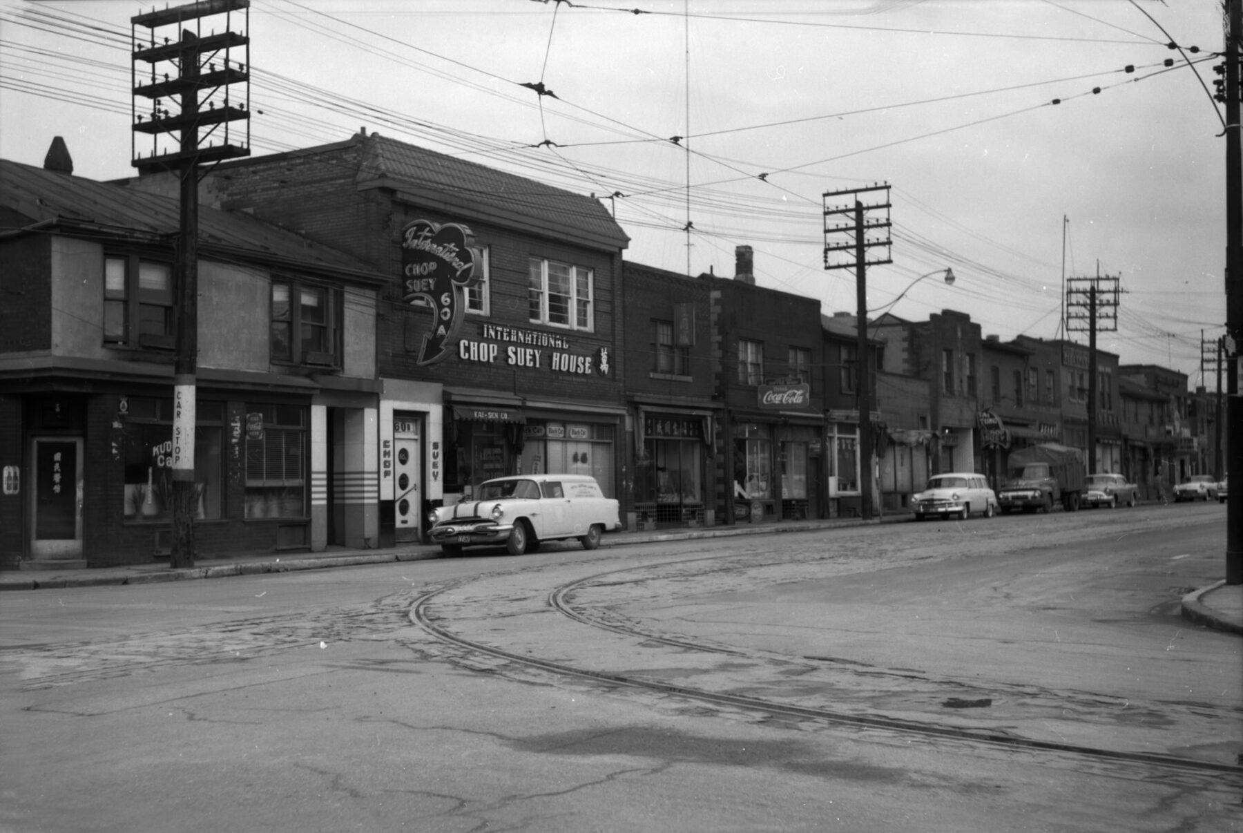 A black and white photograph depicting a street scene. A  series of one and two-storey buildings can be seen behind a row of electric poles. The largest building has a sign out front that reads "International Chop Suey House."