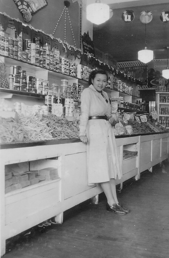 Black and white photograph of a young women in a dress reclining against a display case inside a fruit and grocery store.