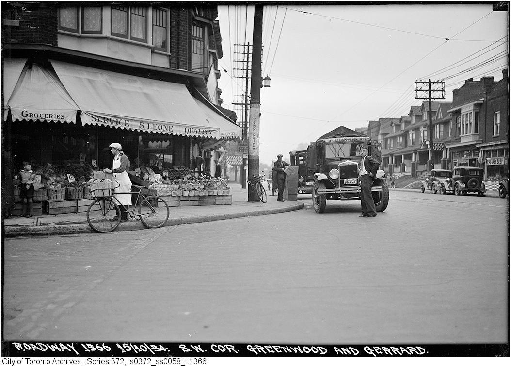 A black and white photo of a grocery shop on the corner of the street, looking west along Gerrard St E. A man pushes a bicycle in front of the shop. Cars are parked along the street and a man stand in front of the nearest vehicle, parked beside the street corner.