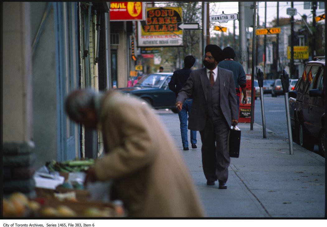 Photo faces east down the sidewalk of Gerrard St E. on the north side. A Sikh man in a suit and carrying a briefcase walks toward the camera. An older gentleman is out of focus in the foreground, selecting vegetables at a store front. The shop sign Sonu Saree Palace and the street sign Hiawatha Rd are visible in the background.