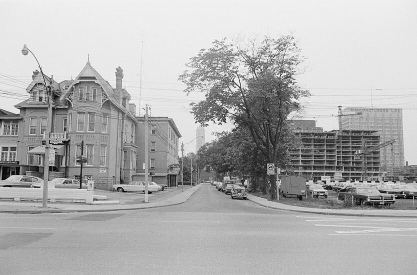 An empty city street, across which a multi-storied Victorian mansion on the left and a modern building under construction on the right can be seen.