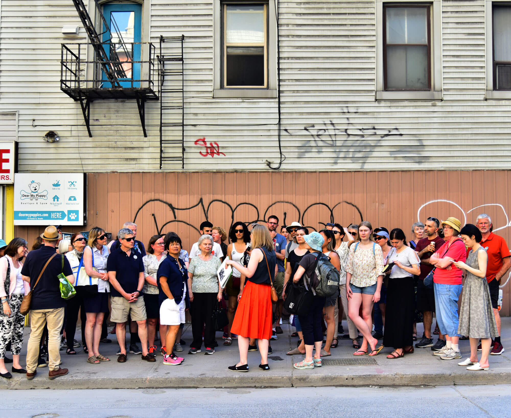 A large group of people stand on a sidewalk. Behind them is a building with some graffiti and storefront signs partially visible, along with a fire escape ladder system.