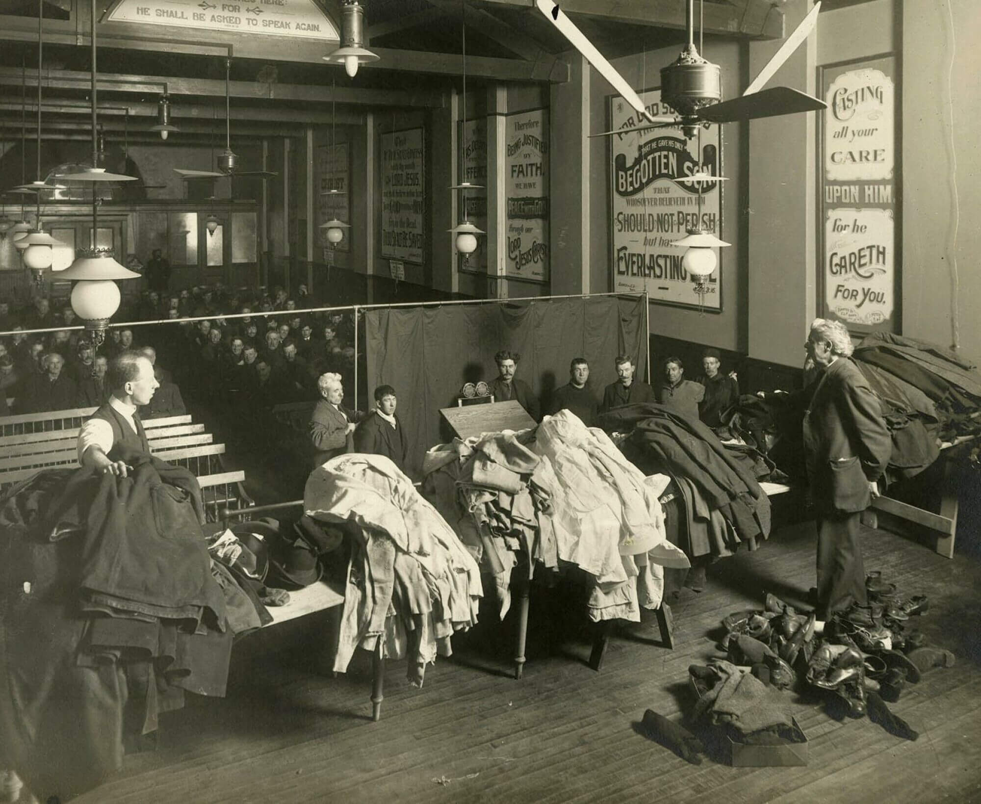 A group of men sitting in a room. In the front is a line of men waiting to go up to an area with clothes. More men are standing with with tables full of clothing (shirts, pants, and shoes).