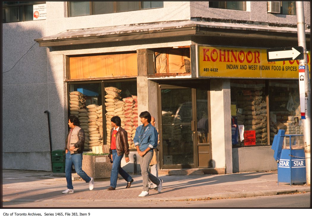 Three young South Asian men walking past a shop with a sign that reads Kohinoor Foods Indian and West Indian Food and Spices.