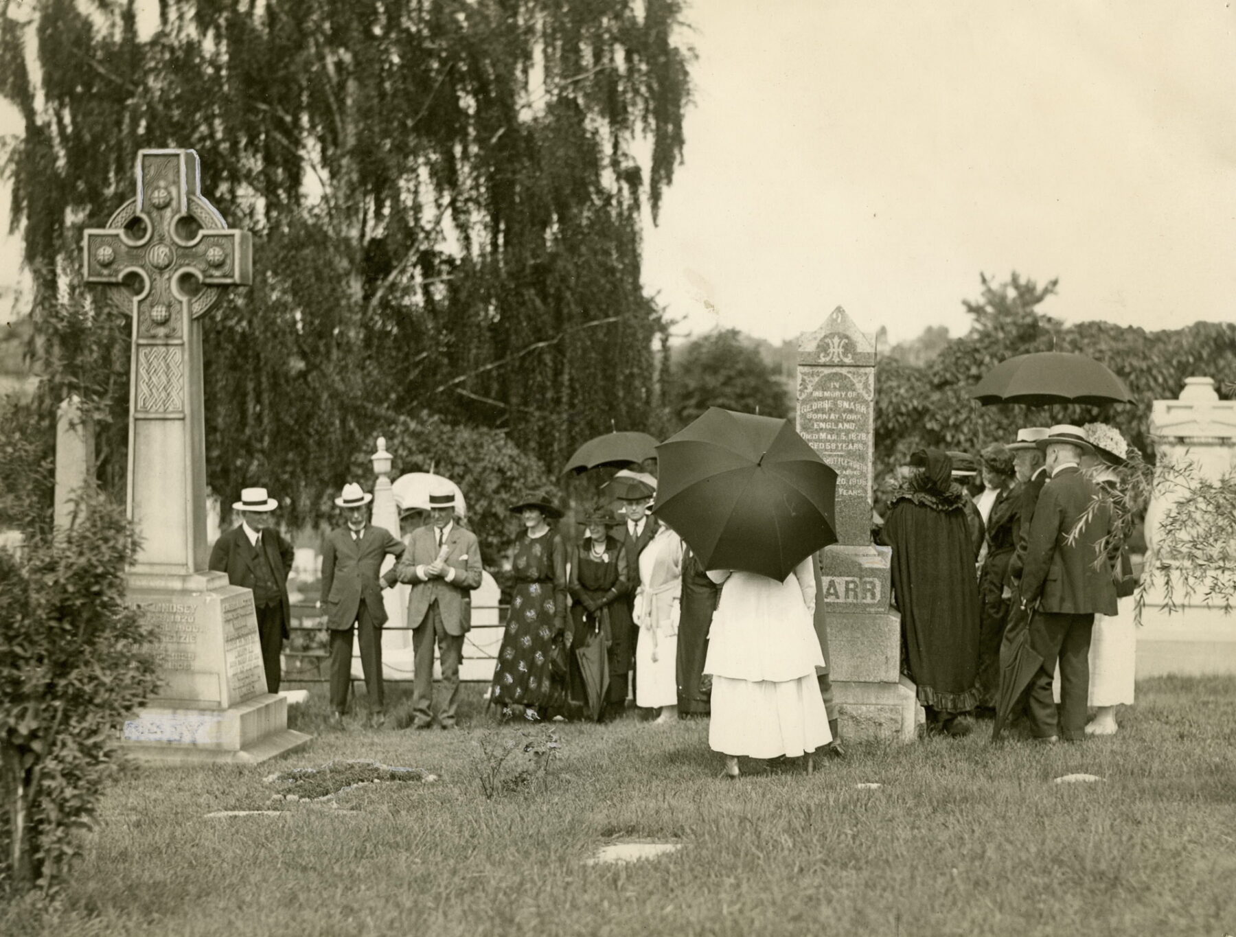 Black and white image of men and women surrounding a grave approximately 12 feet tall topped with a Celtic cross. The people are wearing black and white clothing and some are carrying black umbrellas. Other graves are visible around them.