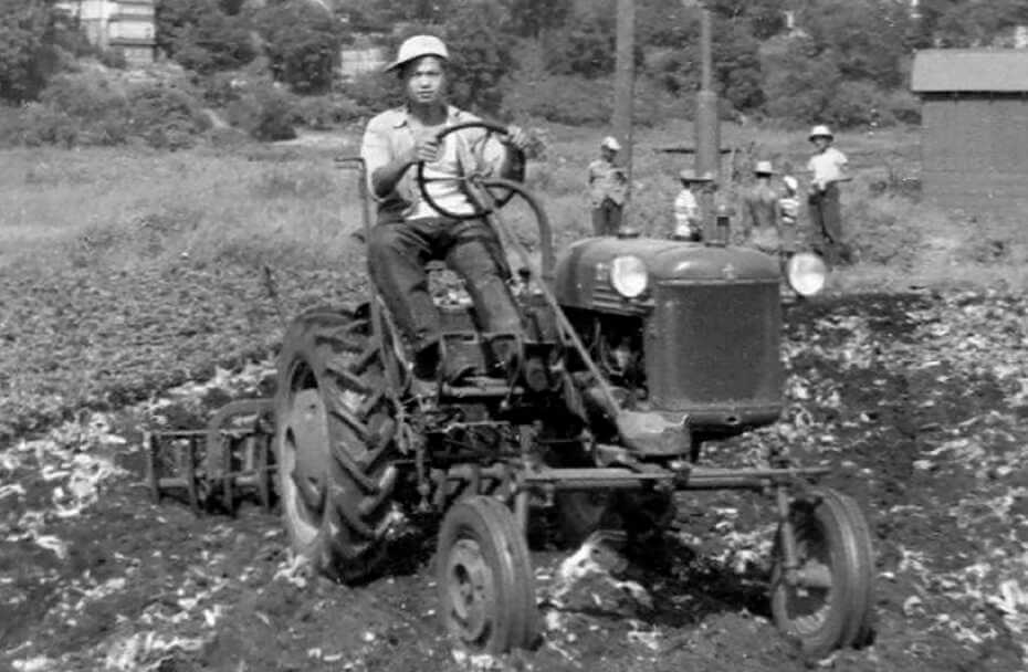 Black and white image of a young man driving a tractor with a plow attached at the rear. A group of men stand together in the background