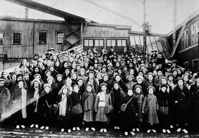 A large group of children stand at a landing dock. They are wearing winter clothing. Many have satchels to carry their belongings.