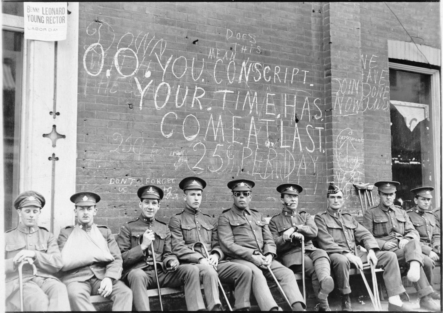 Nine men are seated in a row wearing military uniforms and holding assistive devices including canes and crutches. On the brick wall behind them is graffiti reading, in part, “Conscript your time has come at last,” “Join now,” and “Don’t forget us.”